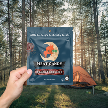 A hand holding two packs of Meat Candy at a woodland campsite