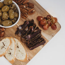 Sliced biltong on a wooden board with olives, pecans, grapes and bread